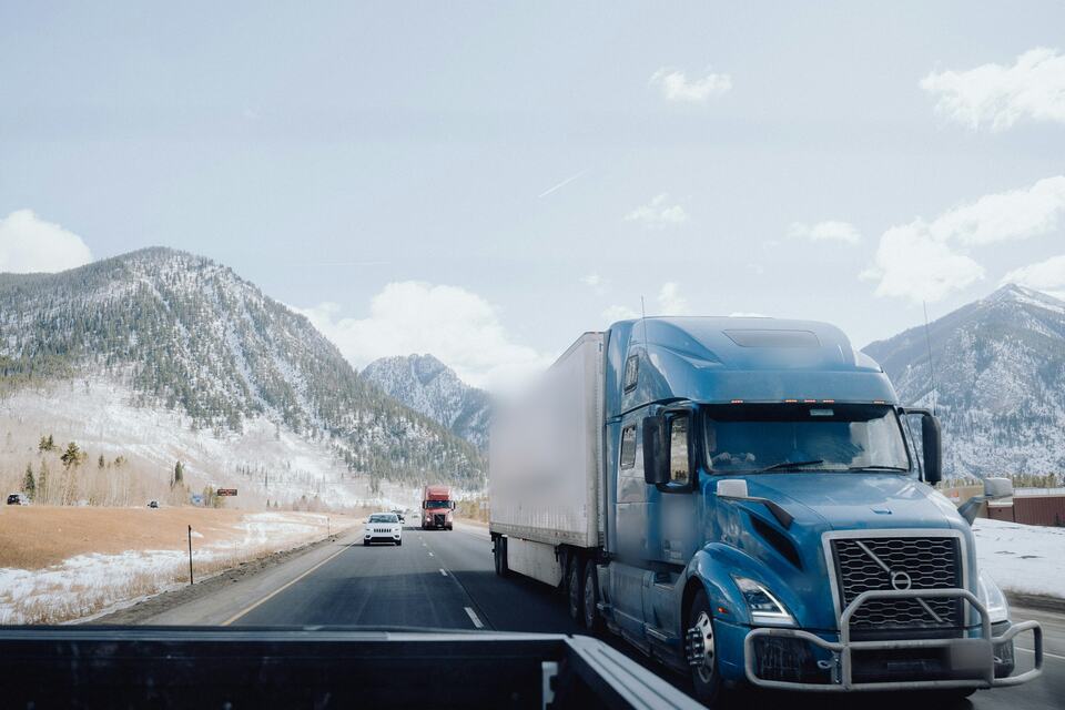 A blue sleeper-cab semi truck pulling a dry van trailer along a snowy mountain interstate